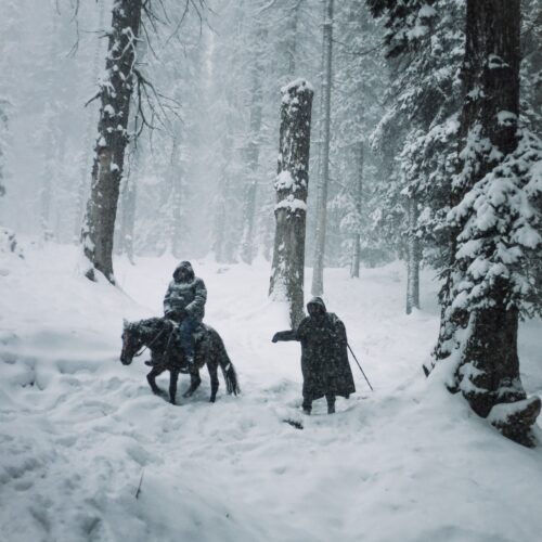 A snowy forest scene featuring a horse, rider, and a companion on a winter trail.